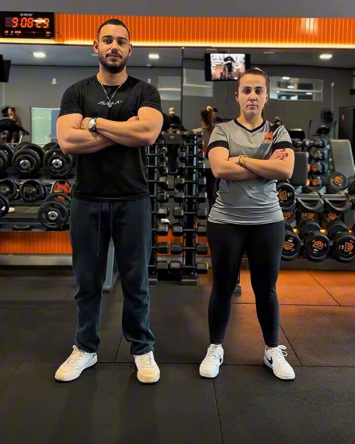 Personal e instrutora da D-Finity Fitness de braços cruzados no salão, com halteres ao fundo