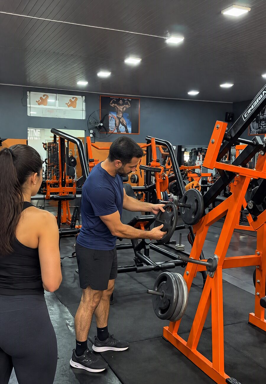 Salão da D-Finity Fitness com aparelhos de musculação laranja e um aluno colocando anilha